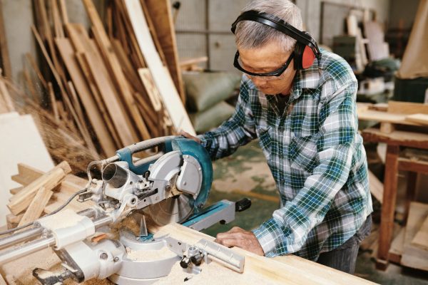 Experienced elderly carpenter in protective goggles and earmuffs cutting wooden planks with circular saw