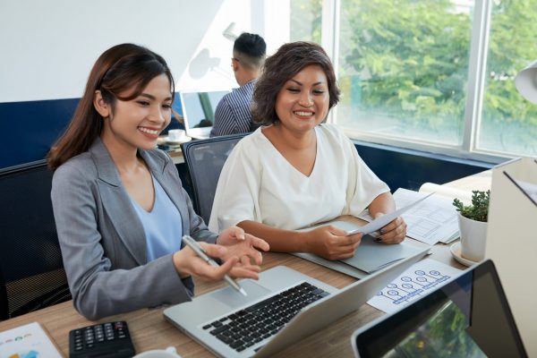 Smiling Vietnamese female coworkers discussing information on laptop screen